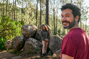 Actor portrayal of an adult man at least 19 years of age smiling in the woods with man and woman sitting on rocks in the background