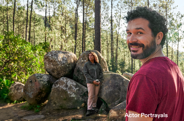 Actor portrayal of an adult man at least 19 years of age smiling in the woods with man and woman sitting on rocks in the background