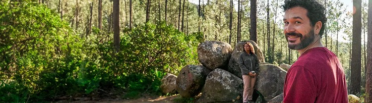 Adult man at least 19 years of age smiling in the woods with man and woman sitting on rocks in the background
