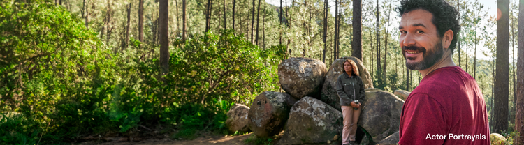 Actor portrayal of an adult man at least 19 years of age smiling in the woods with man and woman sitting on rocks in the background