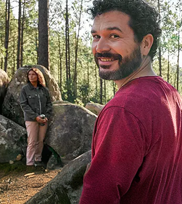 Adult man at least 19 years of age smiling in the woods with man and woman sitting on rocks in the background