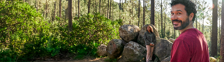 Actor portrayal of an adult man at least 19 years of age smiling in the woods with man and woman sitting on rocks in the background